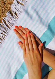 manicured hands with orange nail polish on nails resting on beach towel 
